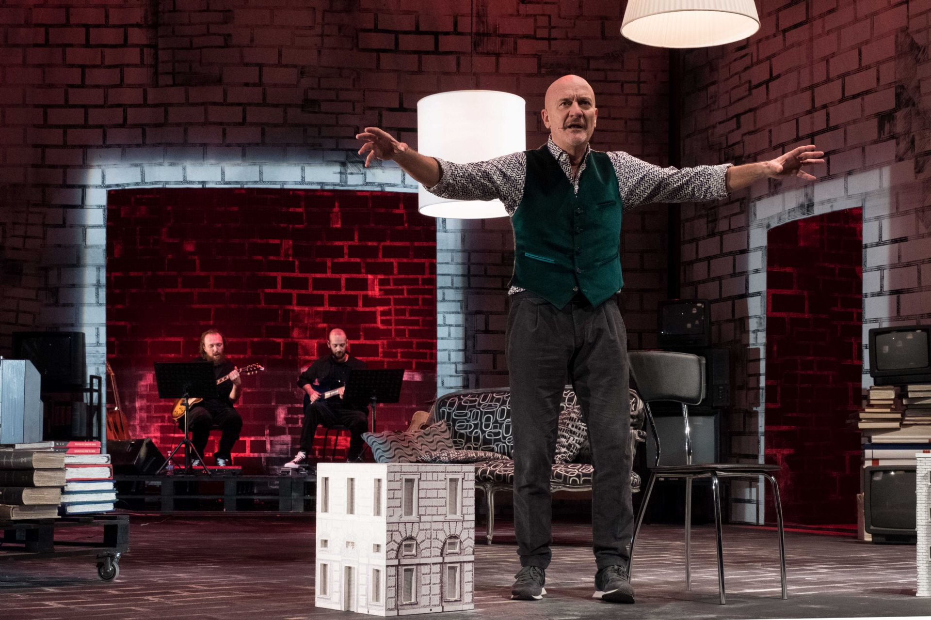 Cludio Bisio all'interno di un salotto di scena con sedie, divani e libri e sullo sfondo due musicisti