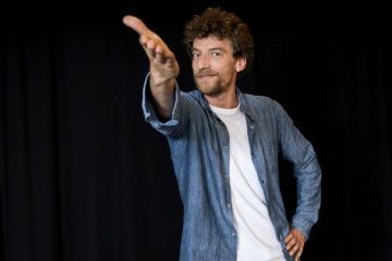 Maurizio Lastrico con sguardo in camera e braccio sollevato e postura rivolta allo spettatore come a dire "guarda te!"
