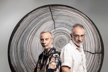 Giovanni Storti e Stefano Mancuso schiena a schiano con lo fondo una riproduzione dei cerchi di un tronco d'albero